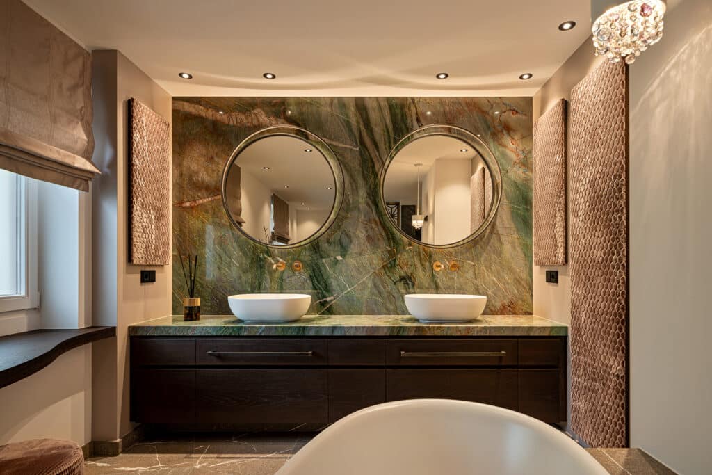 Badezimmer mit Doppelwaschtisch und grün marmorierter Natursteinwand hinter zwei runden Spiegeln