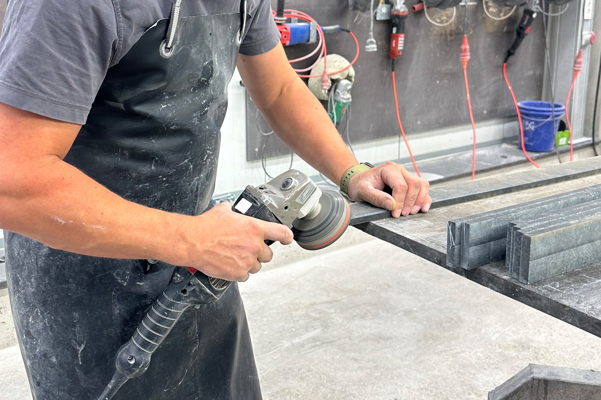 Steinmetz mit Lederschürze bearbeitet die Kante einer Natursteinplatte mit einem Winkelschleifer in der Werkstatt